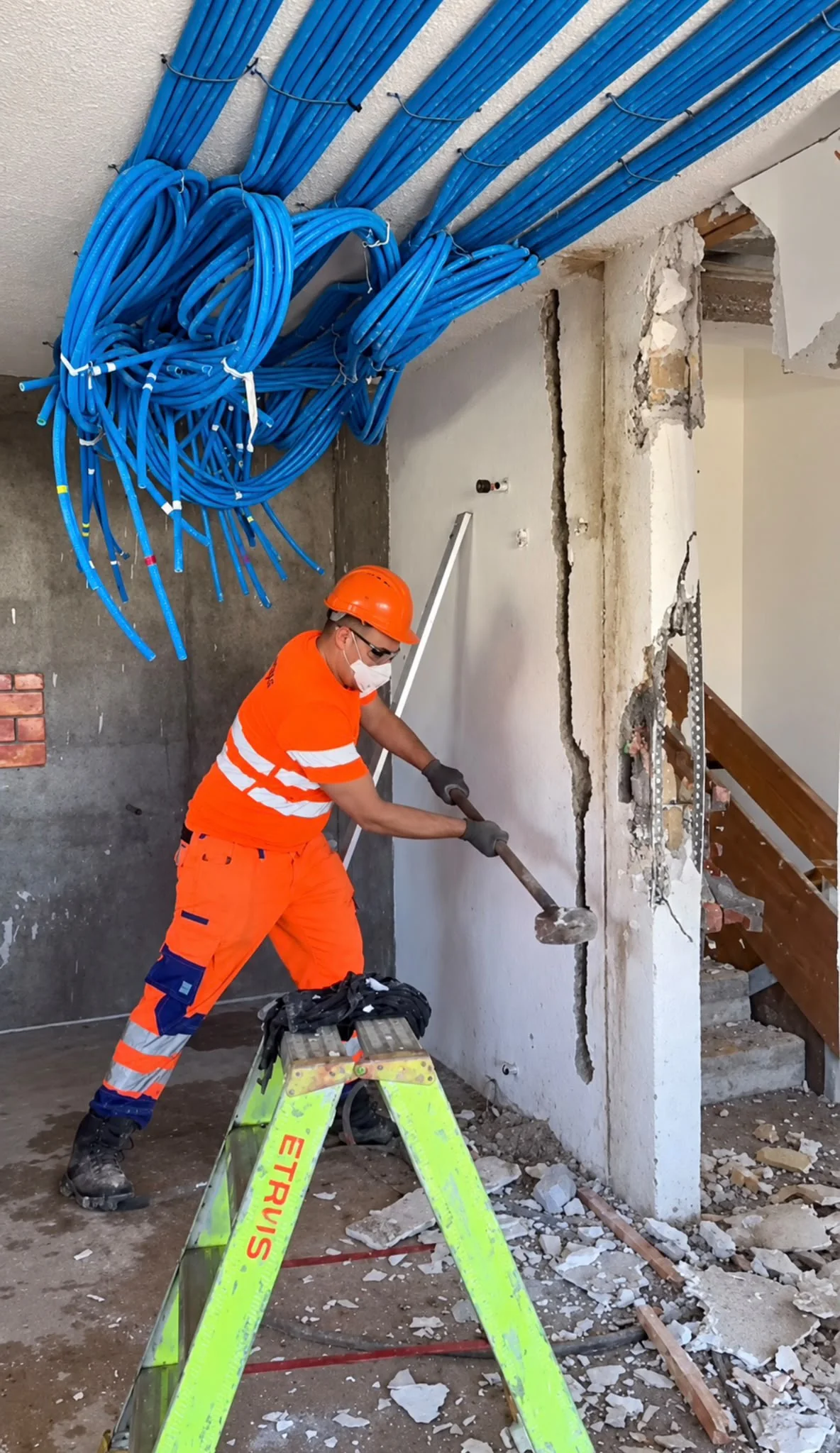 Mitarbeiter in oranger Arbeitskleidung mit Helm und Schutzmaske beim Rückbau einer Wand, an der Decke blaue Leerrohre